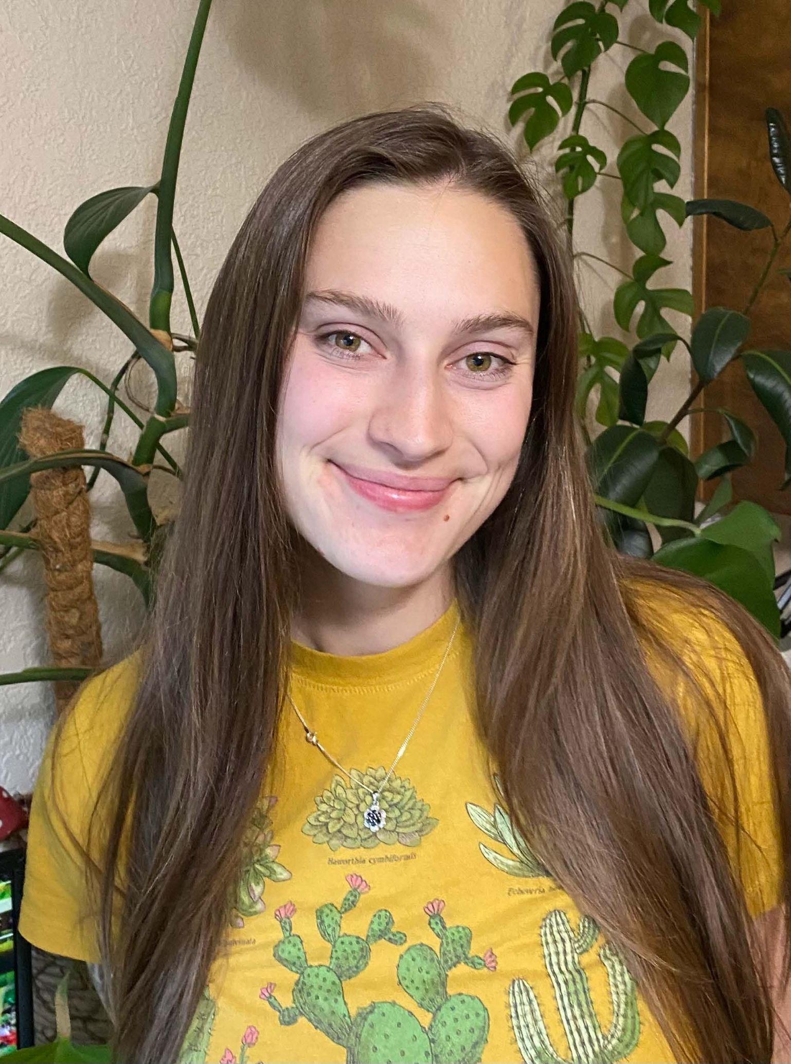 Hannah smiles at the camera wearing a yellow shirt with plants behind her.