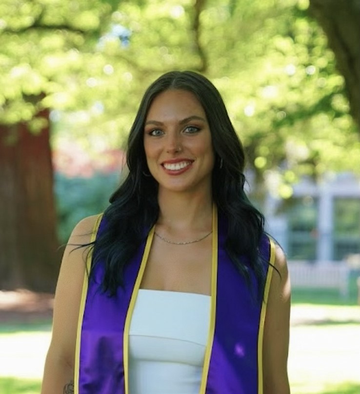 Lila looks at the camera wearing a white dress and a purple graduation sash.
