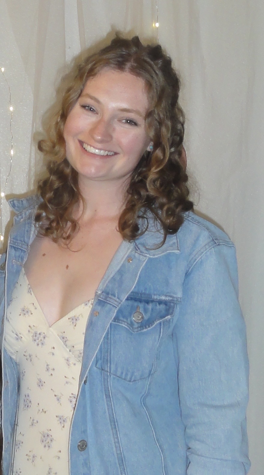 Sophia smiles at the camera wearing a cream colored dress with flowers and a jean jacket.
